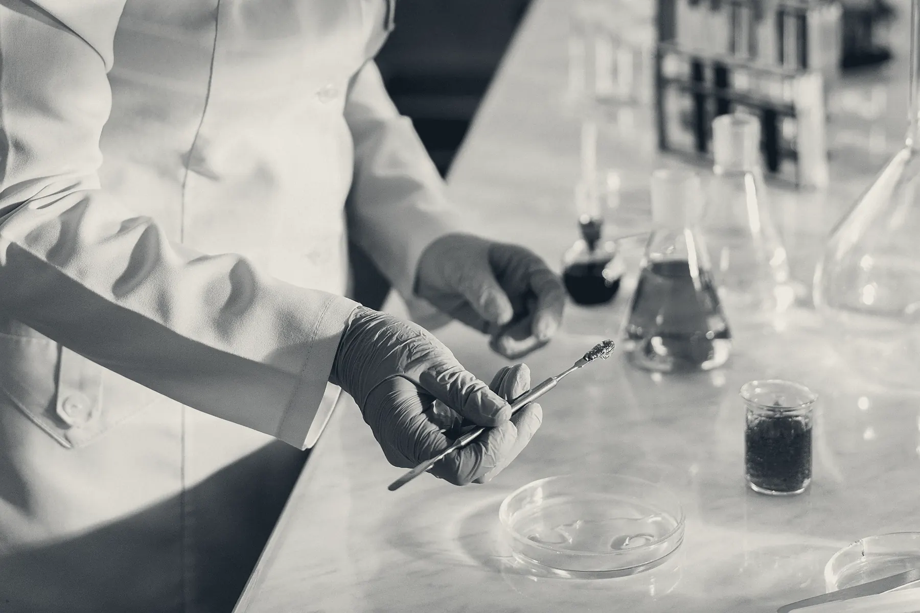 Gloved hands at a laboratory bench dispensing onto sample dishes, glassware on the bench behind. No face and no brand mark.